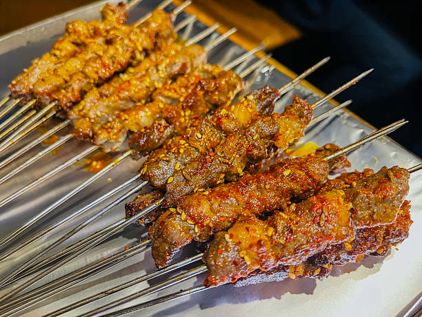 A close-up of several rows of Yang Rou Chuan (羊肉串), a popular and iconic street food originating from the Xinjiang region of China. These mutton or lamb skewers are expertly grilled over charcoal, giving them a distinct charred and smoky aroma. Each skewer consists of small, succulent pieces of meat seasoned with a vibrant blend of traditional spices, most notably toasted cumin seeds, coarse chili flakes, and salt. 

The meat often includes small bits of fat that render during the grilling process, making the skewers incredibly juicy and flavorful. The skewers are typically prepared on long, thin metal needles, as seen in the image, which allow them to be rotated easily over high-heat grills. This dish is a staple of Northeastern Chinese BBQ and is frequently enjoyed as a savory snack or a hearty meal accompanied by cold beer.